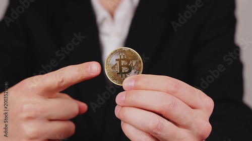 Une main montre une pièce de Bitcoin. En arrière plan, une personne en costume et chemise blanche.
