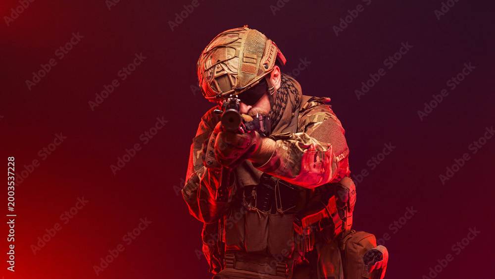 Fototapeta premium Military fighter in tactical gear with rifle on red smoky studio background. Concept combat and defense. 
