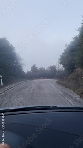 POV driving shot from inside a car moving along a moody foggy asphalt country road surrounded by trees and mist in winter