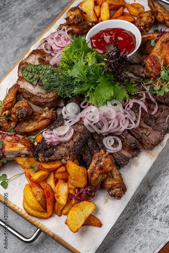 Assorted grilled meats with chicken wings, beef steak slices, roasted potato wedges, fresh onion rings, greens and ketchup sauce served on wooden board
