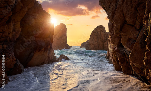 Sand beach among rocks on evening sunset. Ursa Beach near Cape Roca (Cabo da Roca) at Atlantic Ocean coast in Portugal. Summer landscape. Beach with splashes of waves crashing of stone rocks.
