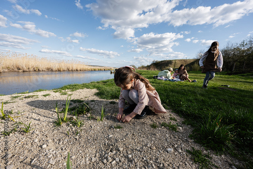 Family picnic by the river with children playing on grass and a little girl exploring the shore on a sunny spring day