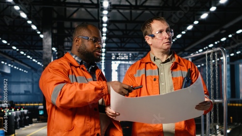 Bild auf Leinwand Two supervisors use plans to oversee construction and production tasks, surrounded by industrial machinery, metal tools and engineering elements that support plant operation