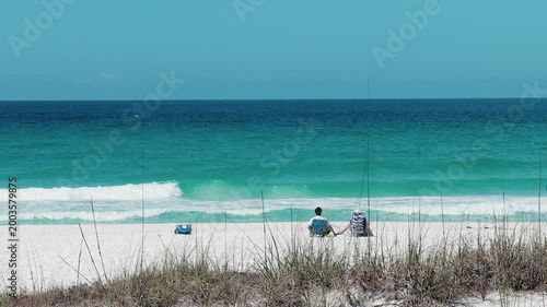 wide shot of couple sitting on white sand beach facing turquoise ocean