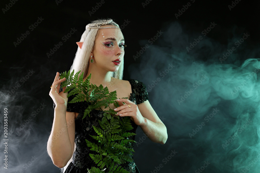 Fototapeta premium Beautiful elf girl with floral decor on black background with smoke, space for text