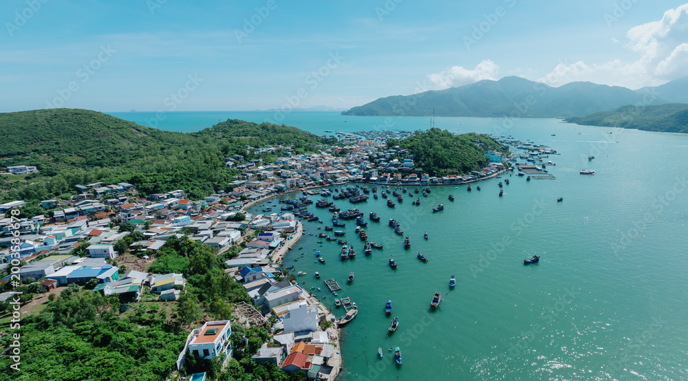 Obraz premium An aerial view of a coastal village in Hon Mieu, Nha Trang, Vietnam, with numerous fishing boats docked in the turquoise bay. The village is nestled between lush green hills and the sea.