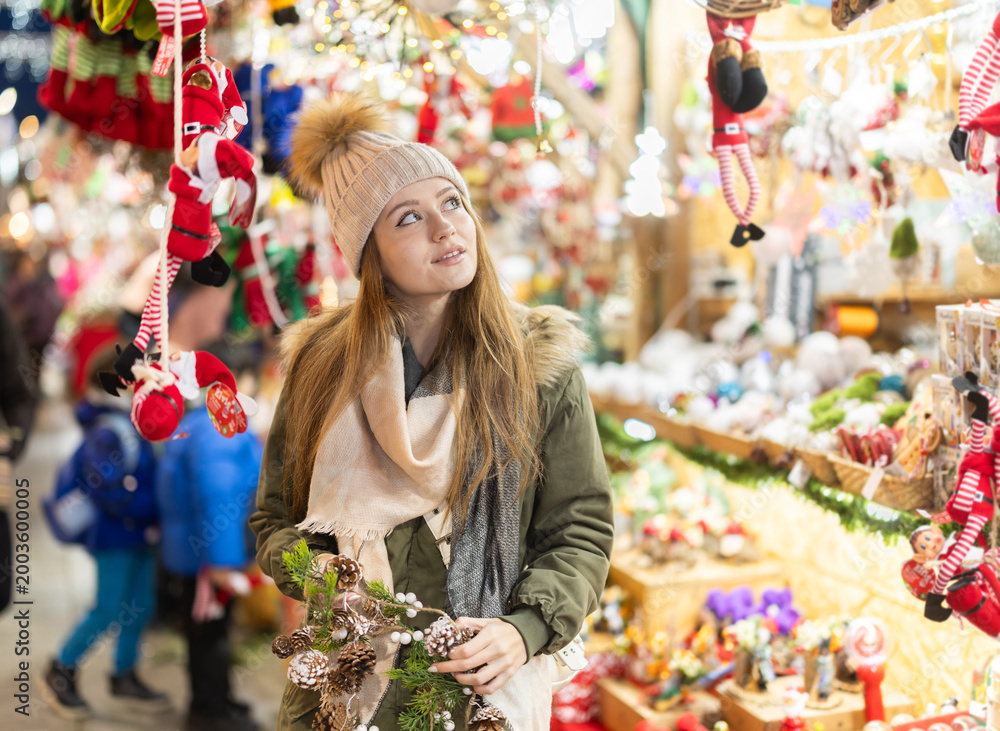 Fototapeta premium Young woman walks view hanging toys near window of New Year souvenirs seller, choose ornaments for Christmas tree. New Years rush. Shes fiddling with garland of fir cones..