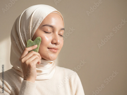 Muslim Woman in Cream Hijab Using Jade Gua Sha Tool for Facial Massage Skincare Beauty Routine