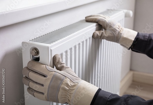 A person wearing work gloves is shown adjusting or maintaining a white heating radiator mounted on a wall.