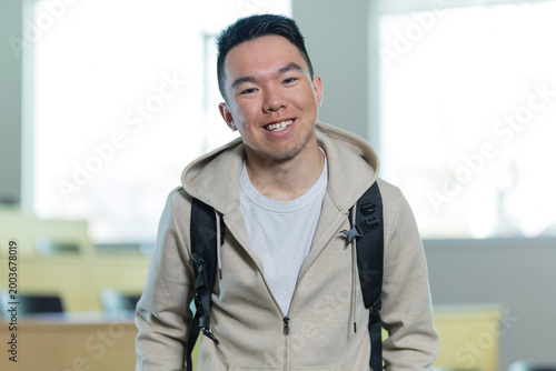 A happy Asian young man smiles directly at the camera while wearing a backpack in a university lecture hall, perfect for higher education, student enrollment, or campus lifestyle concepts.