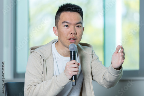 An Asian young man speaks into a handheld microphone while gesturing with his hand in a university lecture hall, perfect for student presentation, public speaking, or campus discussion concepts.