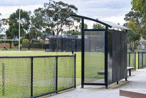Fotografie A metal shelter or sports dugout at the edge of a suburban recreational reserve, overlooking a green sports field with goal posts and children playing in the background