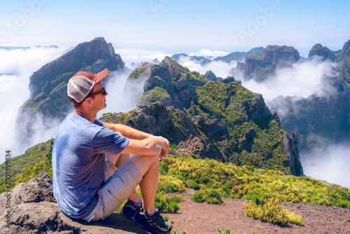 A hiker sits on a rugged mountain peak in Madeira, gazing at a breathtaking landscape of jagged green cliffs and valleys partially veiled by a thick sea of white clouds.