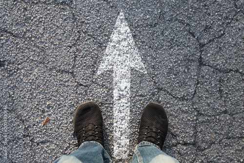 Top view of a person standing on textured asphalt road with a bold white arrow pointing forward.