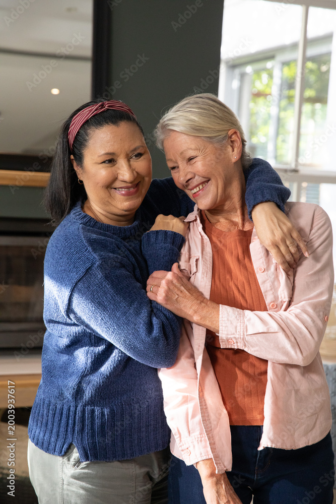 Obraz premium Mature female friends hugging in living room, wearing blue sweater and pink shirt, ring showing