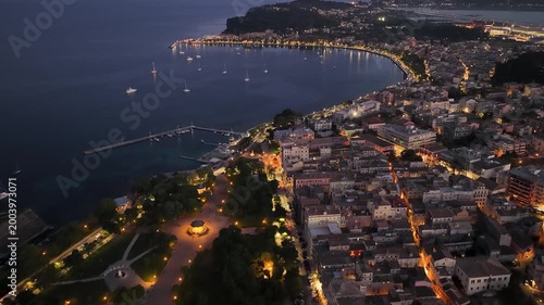 Majestic aerial drone shot of Corfu old town and Spianada Square at dusk, featuring illuminated city streets, historic architecture, and the beautiful curving coastline of Greece