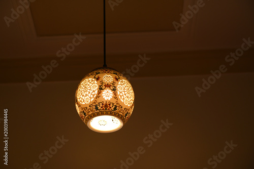 A glowing, decorative pendant lamp featuring intricate floral cutouts and painted patterns, hanging in a dimly lit room.