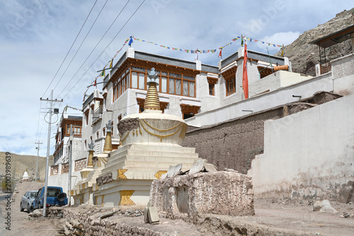 A traditional white building with wooden windows and prayer flags, located behind Buddhist stupas along a mountain road