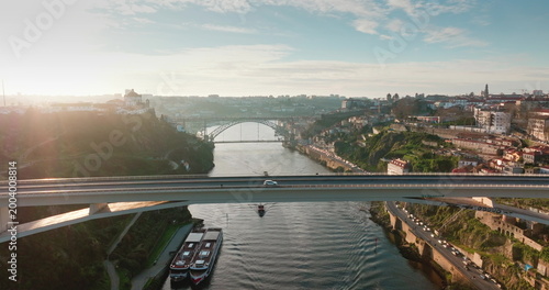 Aerial view flight over bridge in Porto city displaying its historic cityscape, spanning the Douro River, modern Infante Dom Henrique bridge carrying cars driving traffic, against bright sky at sunset