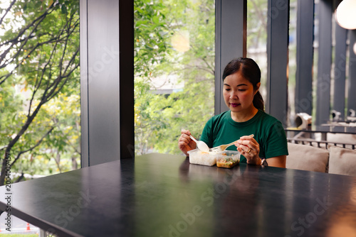 Asian Woman in Green Shirt Eating a Packed Lunch from a Plastic Container in a Modern Cafe with Large Windows