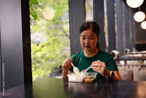 Asian Woman in Green T-shirt Eating Prepared Lunch from Plastic Takeaway Container in Modern Cafe