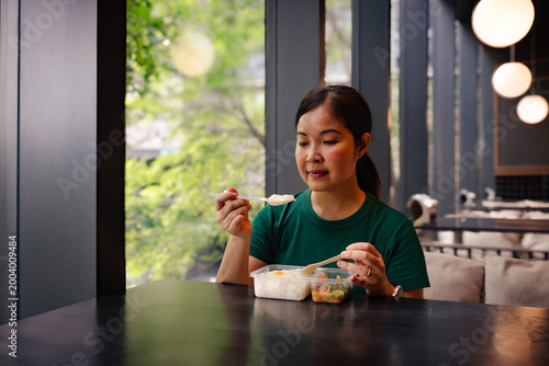 Asian Woman in Green T-shirt Eating Rice with Spoon from Plastic Lunch Box in Modern Cafe