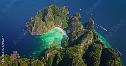 Phi Phi Island Maya Bay aerial: towering limestone cliffs draped in lush green, pristine white sand and vibrant turquoise water with boats cruising coast. Summer vacation paradise, travel to Thailand