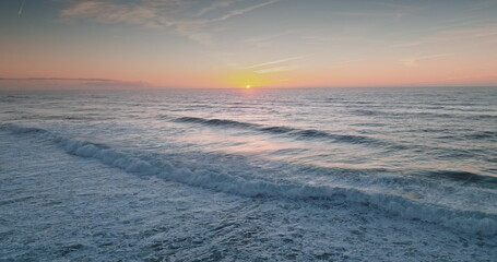 Amazing sunset over the Atlantic Ocean creates pastel colors across the bright sky and reflects on rolling ocean waves. Natural background. Coastal landscape. Aerial drone flight zoom out panorama