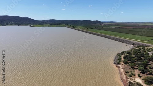 A tracking drone shot of the vast Embalse del Barbate reservoir in Spain, showing the dam wall and surrounding green Andalusian hills under a clear blue sky.