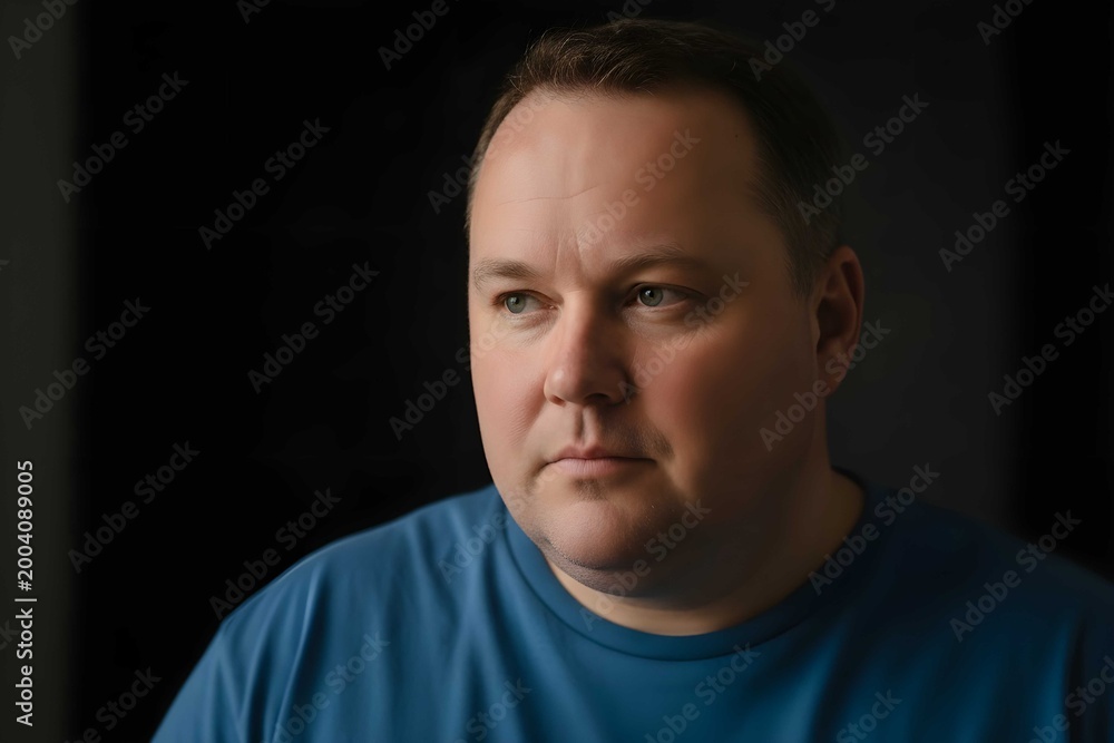 Fototapeta premium Thoughtful overweight man portrait in dark background with soft lighting and serious expression conveying introspection, calm mood and emotional depth