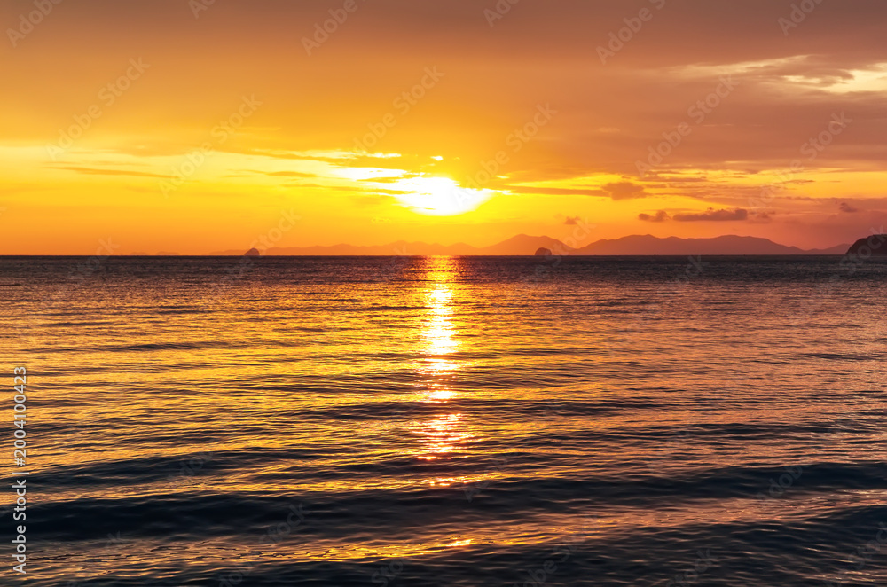 Obraz premium Bright and colorful sunset over the ocean with intense orange tones and shimmering water surface. Ideal tropical travel background with romantic and warm evening atmosphere.