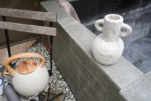 Schilderij op canvas An empty white earthenware clay jar is placed vertically on the corner of a stone fence and modern kettle on wooden chair in an outdoor park