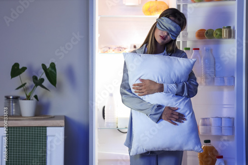 Photos Female sleepwalker with sleeping mask and pillow near fridge in kitchen at night