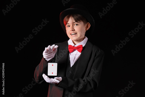 Foto Young magician in tuxedo and top hat performing card trick on black background,