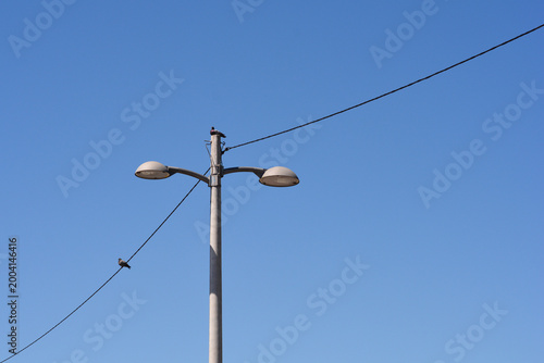 A concrete street lamp with two lights stands under a clear blue sky while two birds perch on the connected overhead power lines