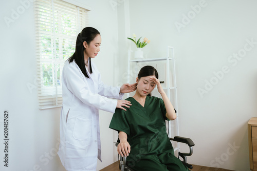 Foto Asian female doctor checks a patient’s condition and offers support as the patient in a wheelchair touches her head, suggesting discomfort in a bright, modern clinic