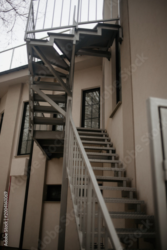 Low angle view of a modern metal fire escape staircase on the exterior wall of a beige building, leading up to a balcony with black window frames on a cloudy day.