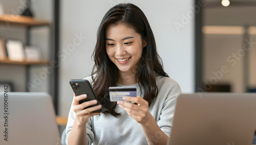 Online Shopping Joy: A young Asian woman with a bright smile is holding her smartphone and credit card, ready to make an online purchase, surrounded by laptops.