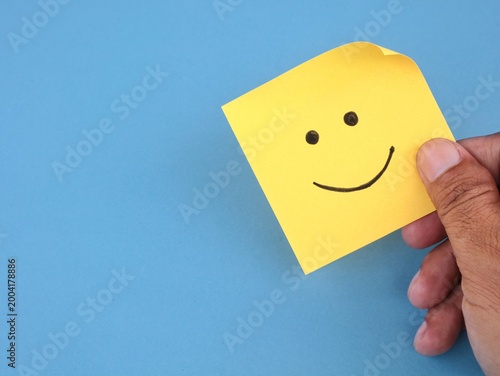 Close-up of hand holding a yellow adhesive note featuring a hand drawn smile face on blue background. Represents customer satisfaction, positive feedback, excellent service rating and happiness 