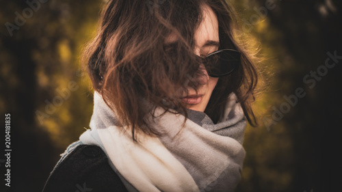 Young woman in sunglasses and scarf standing in a serene autumn setting, partially hidden by her hair, embodying casual elegance and contemplative mood with soft golden tones
