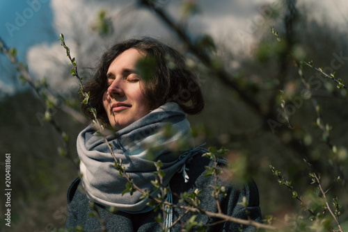 Woman in Cozy Scarf Meditating Peacefully in Nature with Sunlight on Her Face Surrounded by Blurred Branches