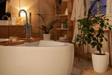 Interior of bathroom with bathtub, plants and glowing lamp in evening