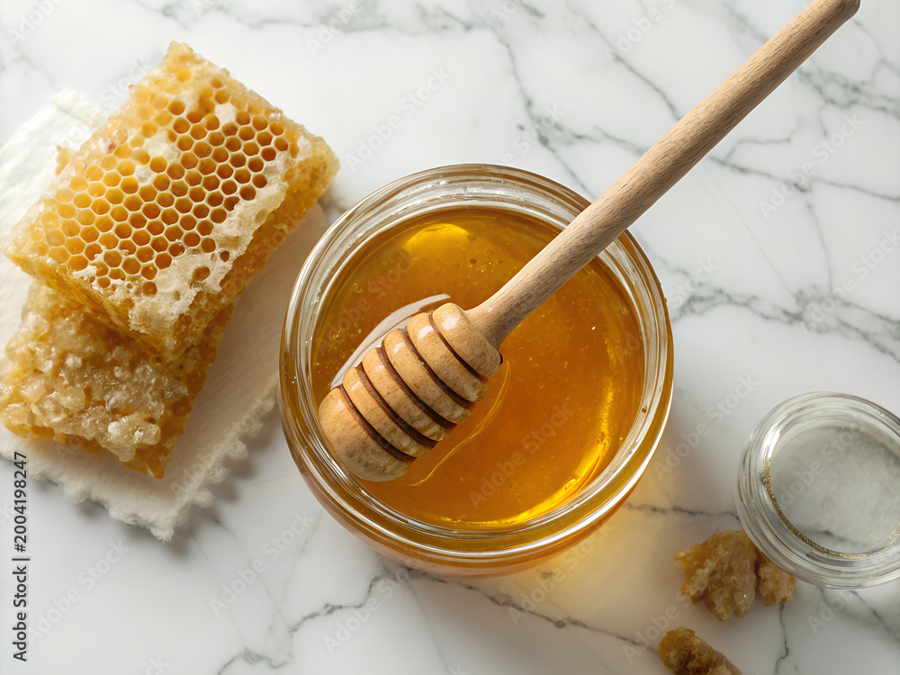 Fototapeta premium Organic raw honey in glass jar with wooden honey dipper and honeycomb on white marble, premium health food photography 4:3 8k 