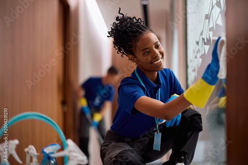 Cuadro en lienzo Happy black female cleaner wiping glass walls in office building.