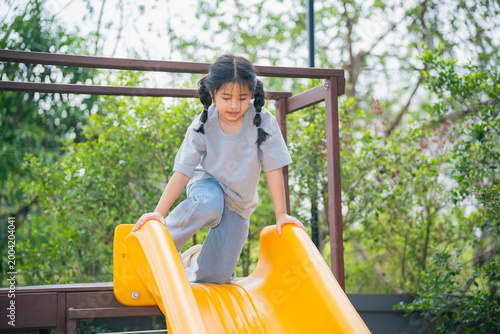 Happy child playing on yellow slide in outdoor playground, enjoying sunny day with trees and greenery in background, carefree childhood fun and laughter