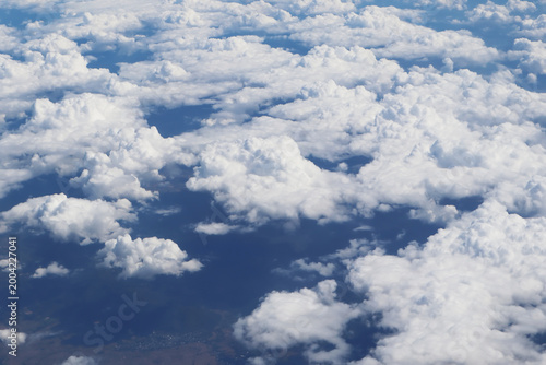 A high-angle aerial photograph taken from an aircraft, showing a vast field of white, fluffy cumulus clouds. The clouds are scattered across the frame, revealing glimpses of the dark blue-toned earth.