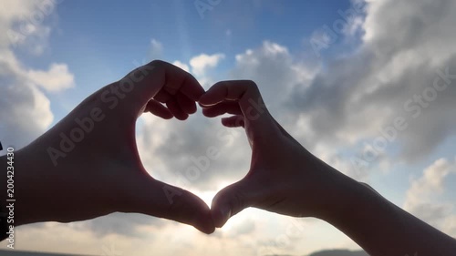 Two hands forming a heart shape around the sun against a beautiful cloudy sky. Concept of love, friendship, and togetherness during sunset at the beach. 4K slow-motion footage.