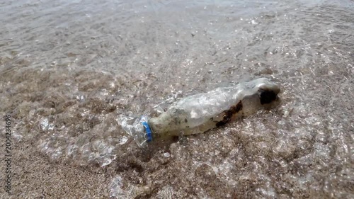 Dirty plastic bottle washed up on the sandy beach by waves. Concept of ocean pollution, and environmental conservation. 4K horizontal slow-motion footage.
