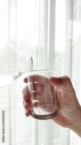 Hand pouring fresh pure water from a plastic bottle into a glass against a bright window background with natural light. Concept of healthy lifestyle, hydration, and clean drinking water.