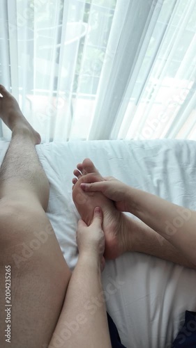 Top view of young man massaging his own feet on a white bed, representing plantar fasciitis pain, basic self-care, and restorative atmosphere for recovery at home. slow motion 4K resolution, vertical.